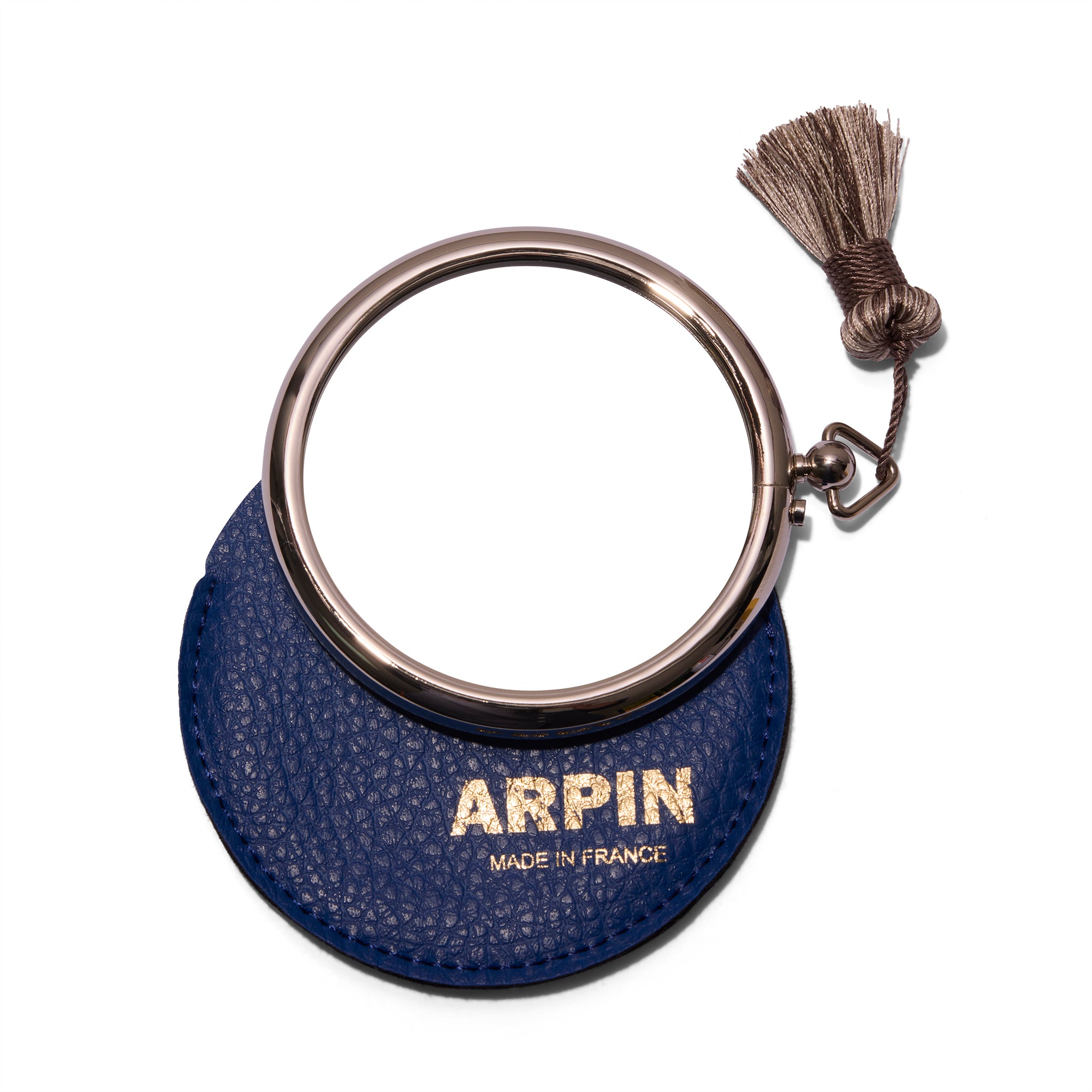 Arpin Hand Bag Mirror from the front and litting on top of the deep blue leather case with gold lettering that says Arpin. The silver grey tassel is on the top right of the mirror.