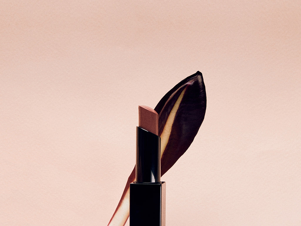 Image of a lipstick in front of a leaf. The background is pink.