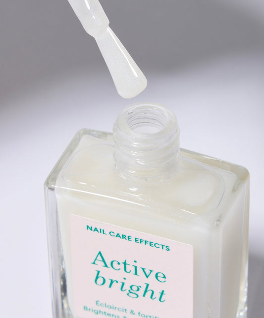 A clear glass nail polish bottle with a sticker with green text. The bottle has a white cap and the polish inside is a cream color with an opal tint. The bottle is open and the brush is outside of the bottle showing the color on the brush.