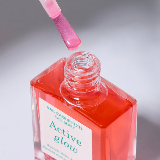 A clear bottle of nail polish with label with green text and a white cap. The polish color is a sheer, light raspberry pink. Cap is off and the brush is just above the opening of the bottle.