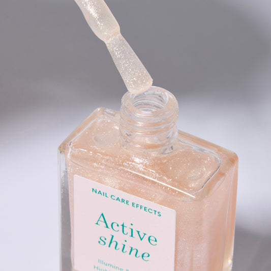 A clear bottle of nail polish with label with green text and a white cap. The polish color is a sheer nude with very soft sparkle. The cap is off and he brush is over the opening of the bottle.