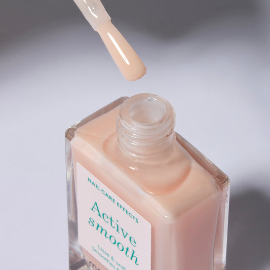 A clear glass nail polish bottle with a white label with green text. The polish is a sheer, soft peach color. The cap is off and the brush is above the opening.