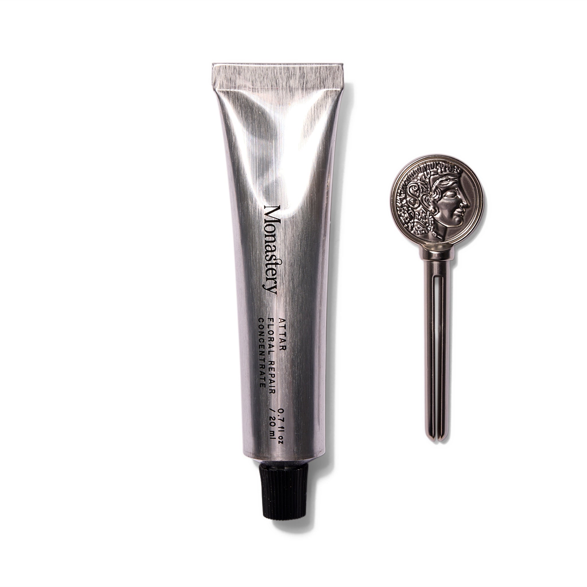 An upright brushed silver tube with a black cap that says Monastery Attar Floral Repair Concentrate. To the right of the tube is a silver key used to squeeze product out. The key has a profile of the Greek goddess Athena.