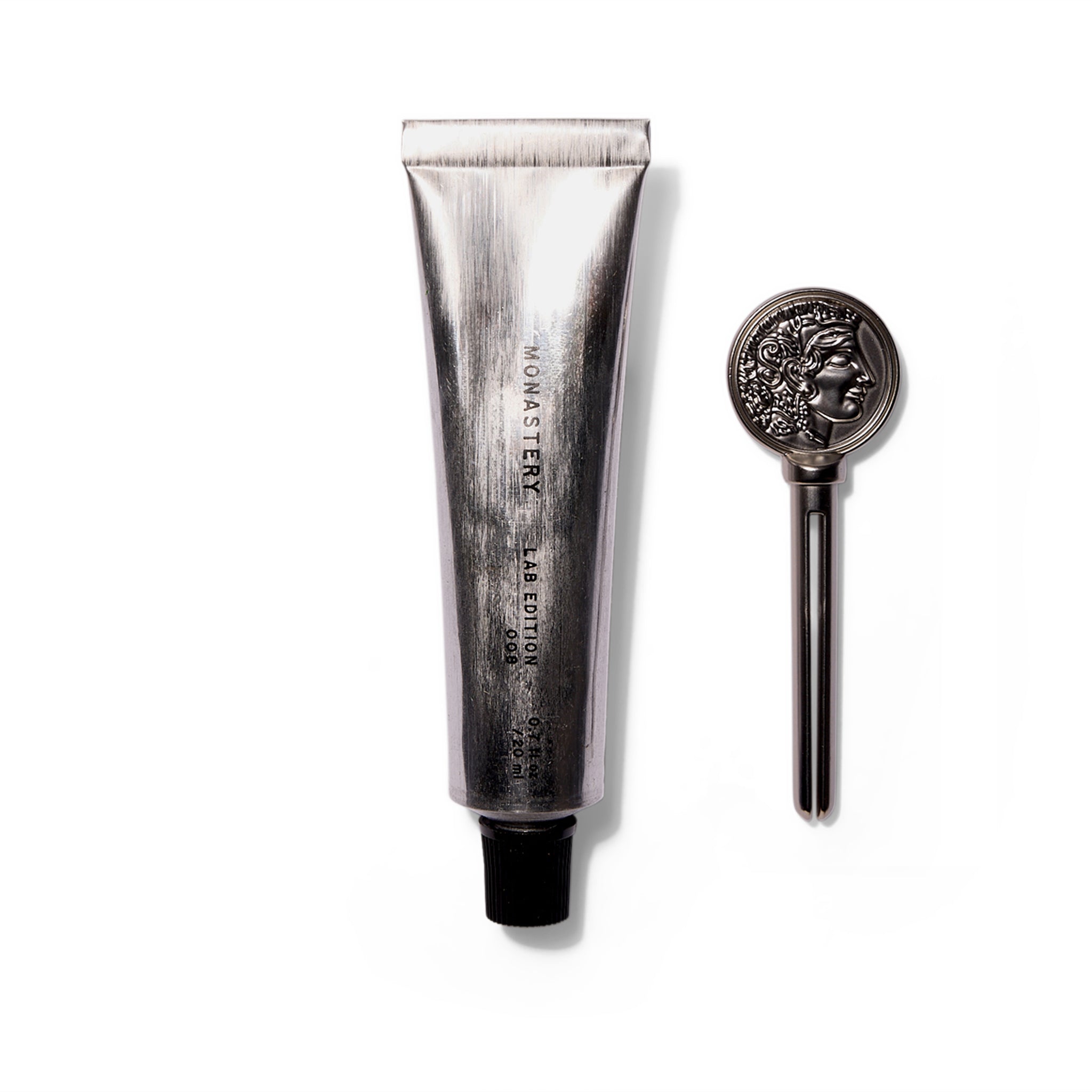 An upright brushed silver tube with a black cap that says Monastery Lab Edition 008 Universal Balm. To the right of the tube is a silver key used to squeeze product out. The key has a profile of the Greek goddess Athena.