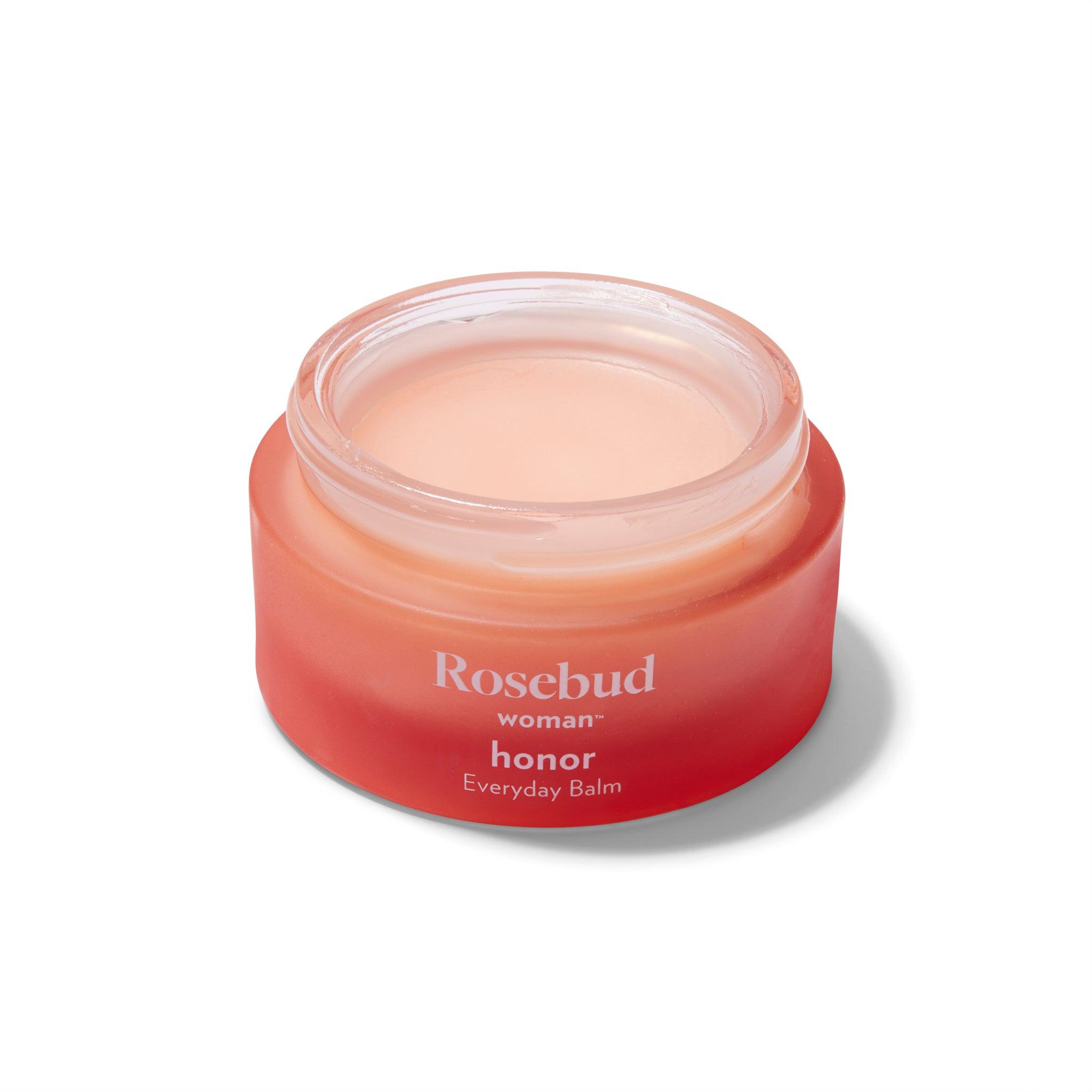 Slighly overhead view of the Rosebud Woman Honor Balm. The jar is a rosy smoked glass and the lid is off showing the pale pink creamy product inside the jar.