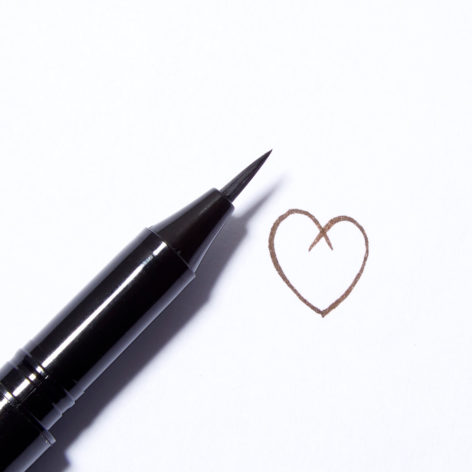  A close up of the Surratt Autographique Liquid Eyeliner in Brun Riche, a rich brown. The pen is made of individual hairs and goes to a sharp point. The pen is angled and there is a heart drawn to the right of it in the ink from the eyeliner.
