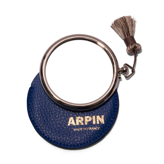 Arpin Hand Bag Mirror from the front and litting on top of the deep blue leather case with gold lettering that says Arpin. The  silver grey tassel is on the top right of the mirror.