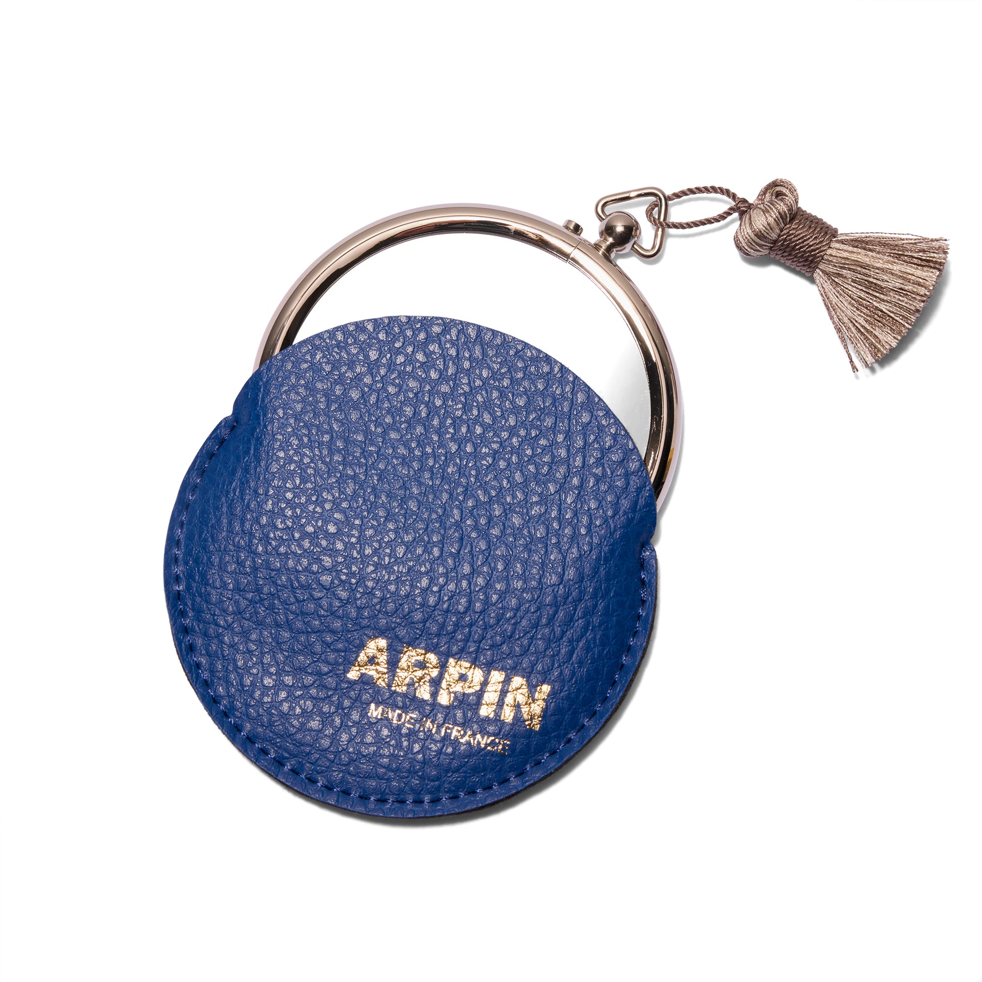 Arpin Hand Bag Mirror from the front and is peeking out of the deep blue leather case with gold lettering that says Arpin. The  silver grey tassel is on the top right of the mirror.