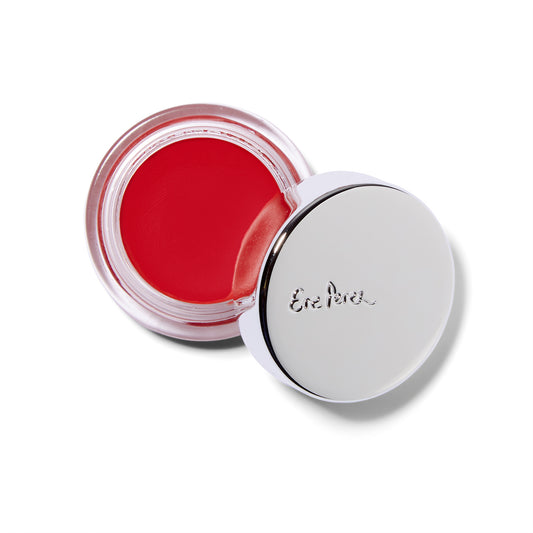 An overhead view of the Ere Perez Carrot Color Pot cream blush. The product is in a squat glass jar with a silver lid. The lid is partially resting on the edge of the glass container. The blush shown is Hello, a bright orange color.