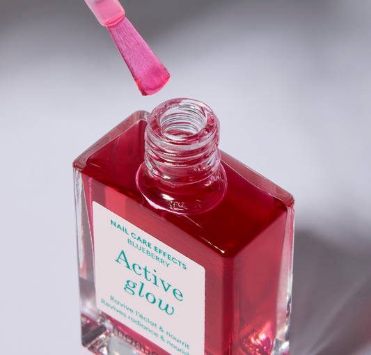 A clear bottle of nail polish with label with green text and a white cap. The polish color is a sheer, cool-toned pink. Cap is off and the brush is just above the opening of the bottle.