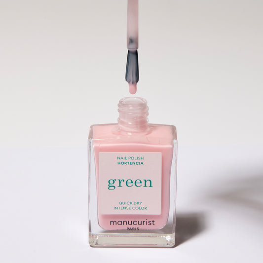 Pale pink nail polish with a white label with green text. The brush is directly over the bottle with a drip of polish on the brush.