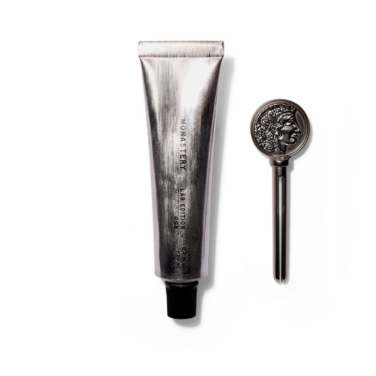 An upright brushed silver tube with a black cap that says Monastery Lab Edition 008 Universal Balm. To the right of the tube is a silver key used to squeeze product out. The key has a profile of the Greek goddess Athena. 